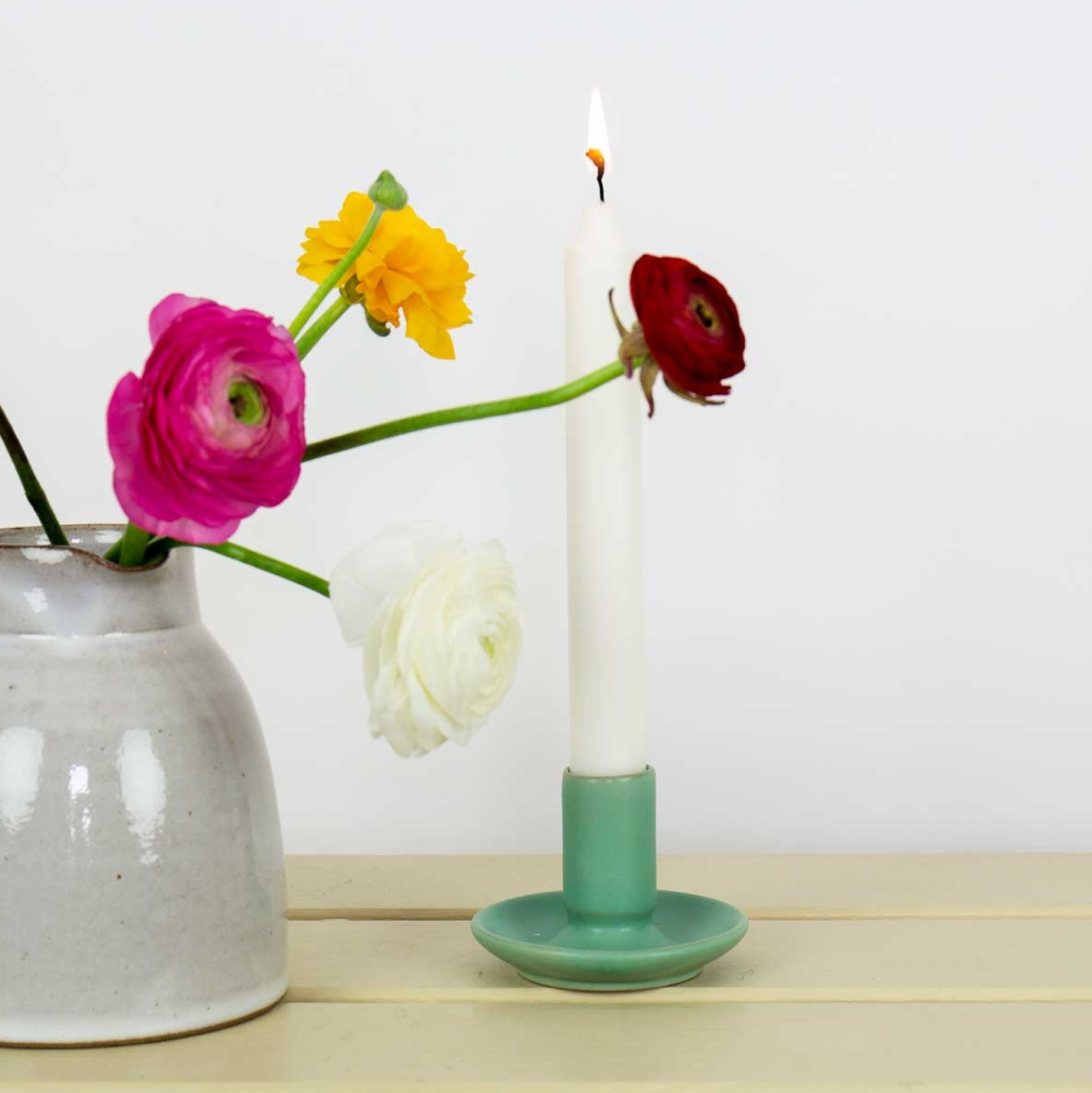 A white candle in the Hera Handmade Candle Holder in green by A World of Craft is surrounded by pink, yellow, red, and white ranunculus flowers next to a ceramic vase on a light wooden surface against a plain white background.