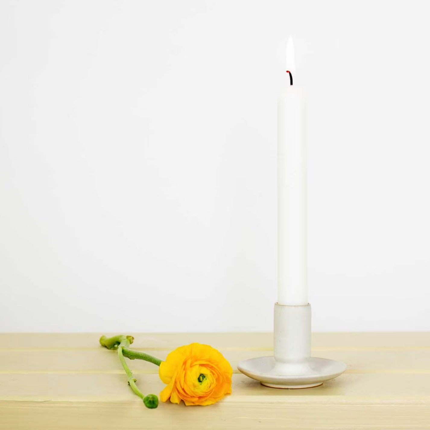 A lit white candle in the Hera Handmade in Beige Candle Holder from A World of Craft stands on a light surface beside a yellow ranunculus with green stem, set against a plain white background.