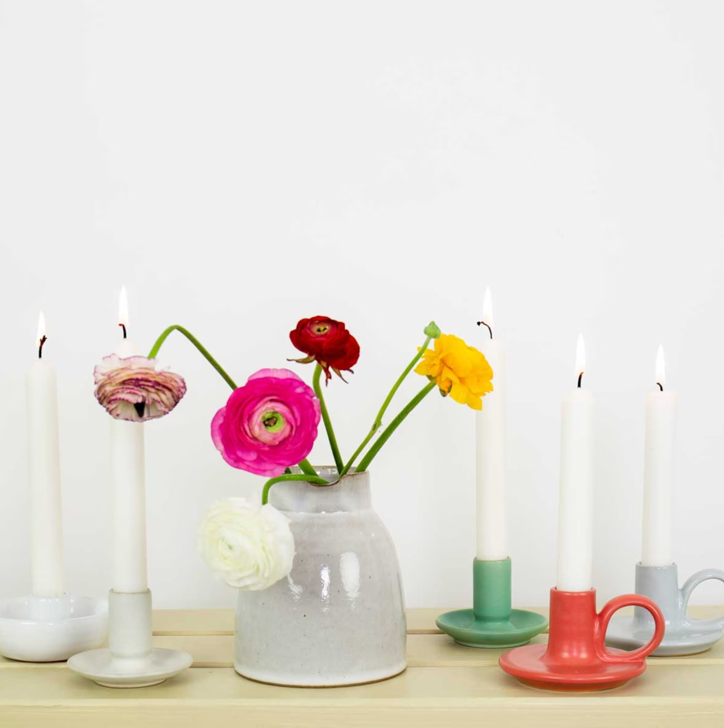 A white vase with colorful ranunculus flowers is surrounded by six lit white taper candles in handcrafted stoneware, including the green Hera Handmade Candle Holder by A World of Craft, on a light wood surface.