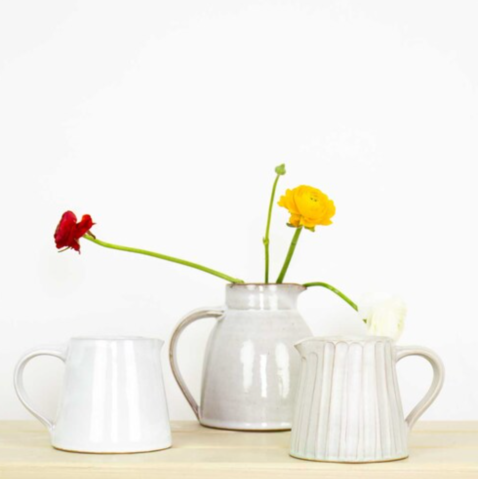 The Rhea Ceramic Jug in beige by A World of Craft sits between two white vessels on a light wood surface, holding one red, one yellow, and one white flower, all set against a plain white background.