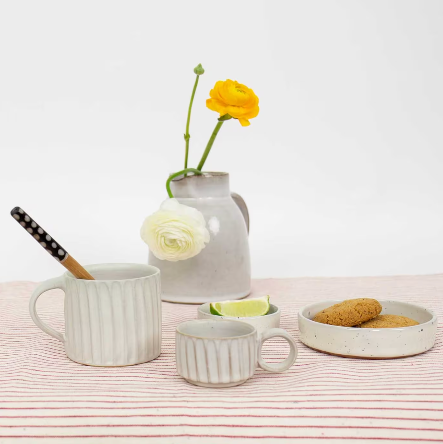 A beige Mini Rhea Handmade Espresso Cup by A World of Craft, a white ceramic mug with a spoon, cookies on a plate, and a jug of yellow and white flowers are displayed on a red-striped cloth against a white background.