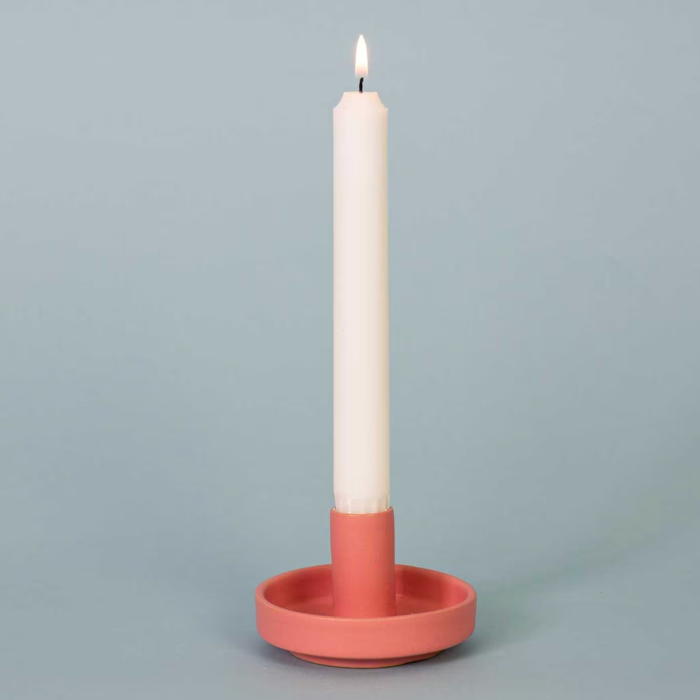 A lit white candle is displayed upright in the Hermes Handmade Candle Holder, Pink by A World of Craft, handcrafted from stoneware and set against a plain light blue background.