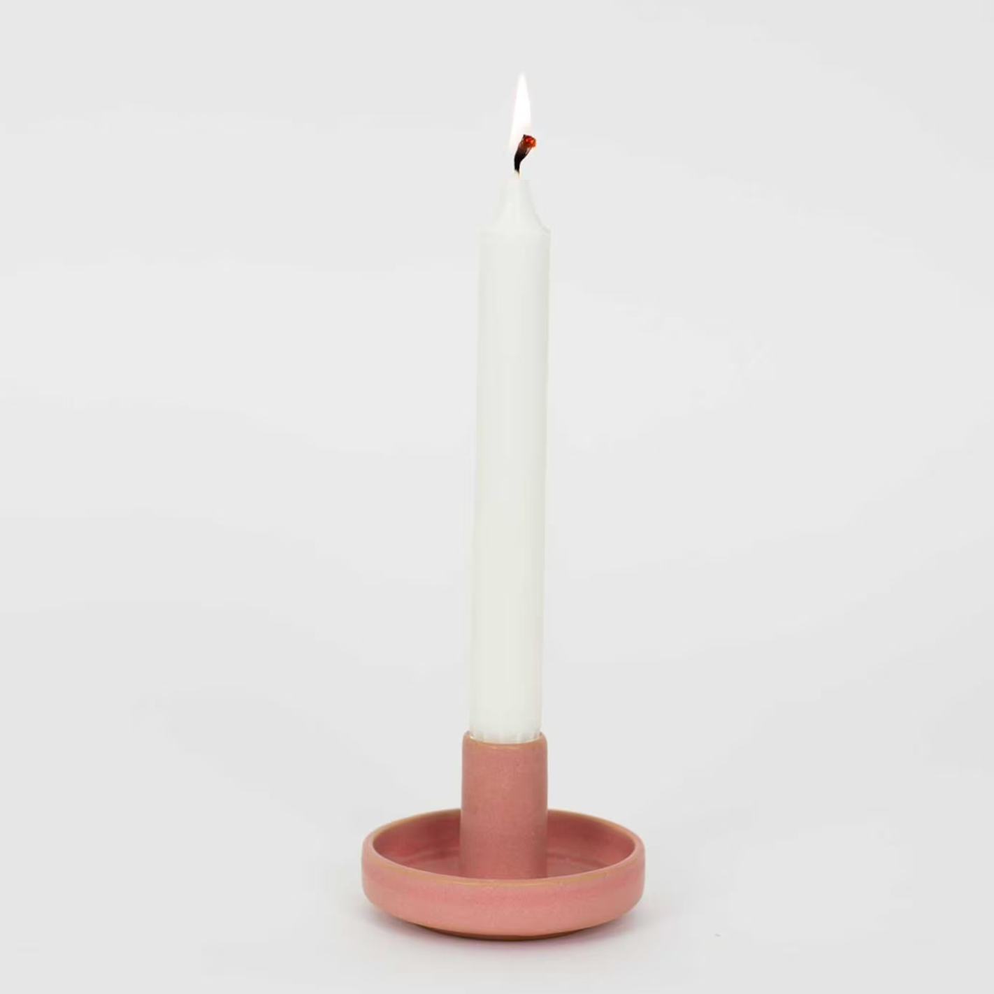 A lit white taper candle sits in the Hermes Handmade Candle Holder by A World of Craft, round and pink stoneware, against a plain white background.