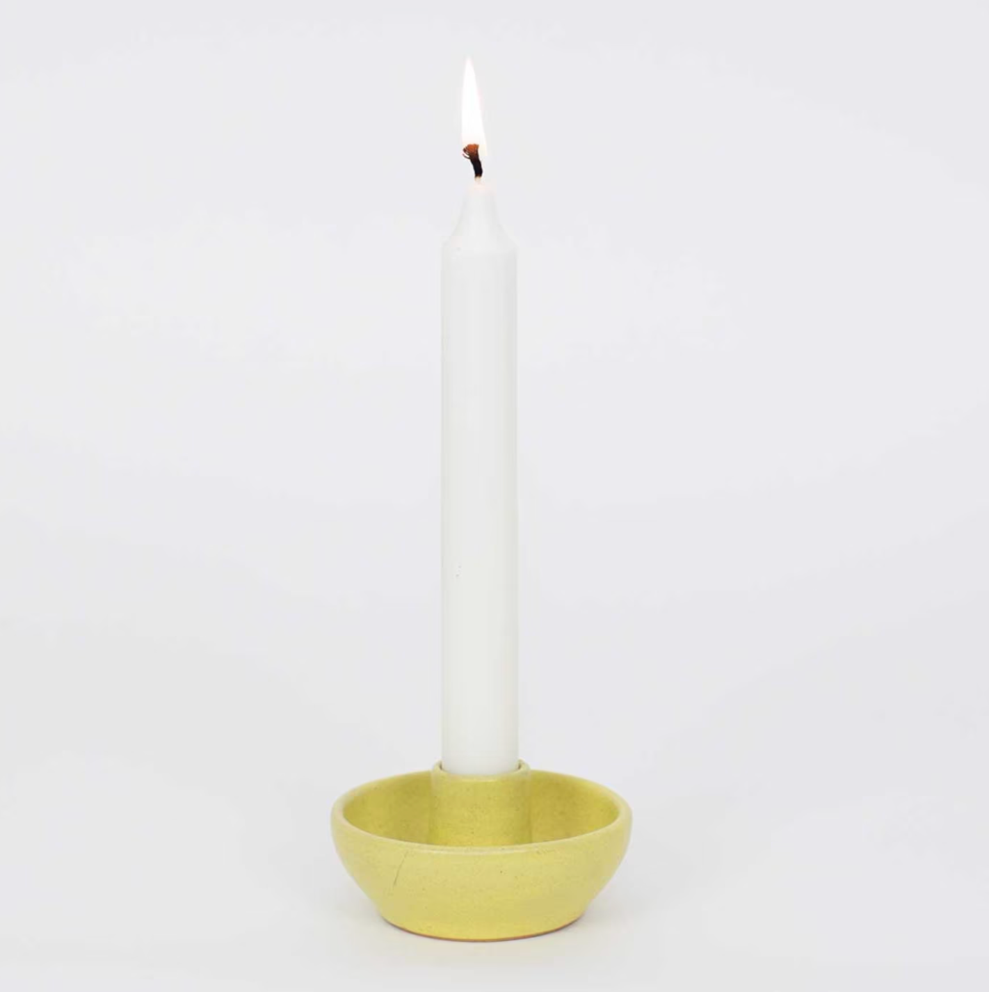 A lit white taper candle stands upright in the Selma Candle Holder, Yellow by A World of Craft, set against a plain white background.