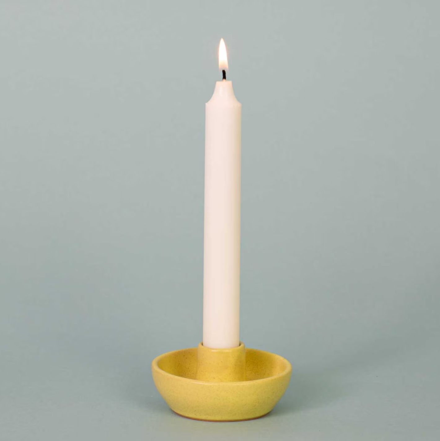 A lit white candle stands upright in the Selma Candle Holder, Yellow by A World of Craft, a small round handcrafted stoneware piece, set against a plain light gray background.