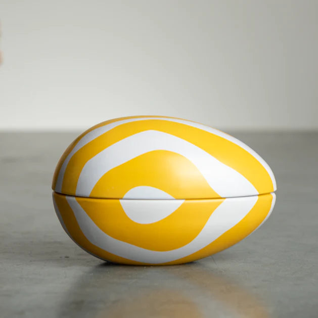 A closed Zebran Reusable Easter Egg by Gåva Gifting, a yellow and white Swedish-style Påskägg tin with wavy concentric patterns, sits on a smooth gray surface against a plain background.