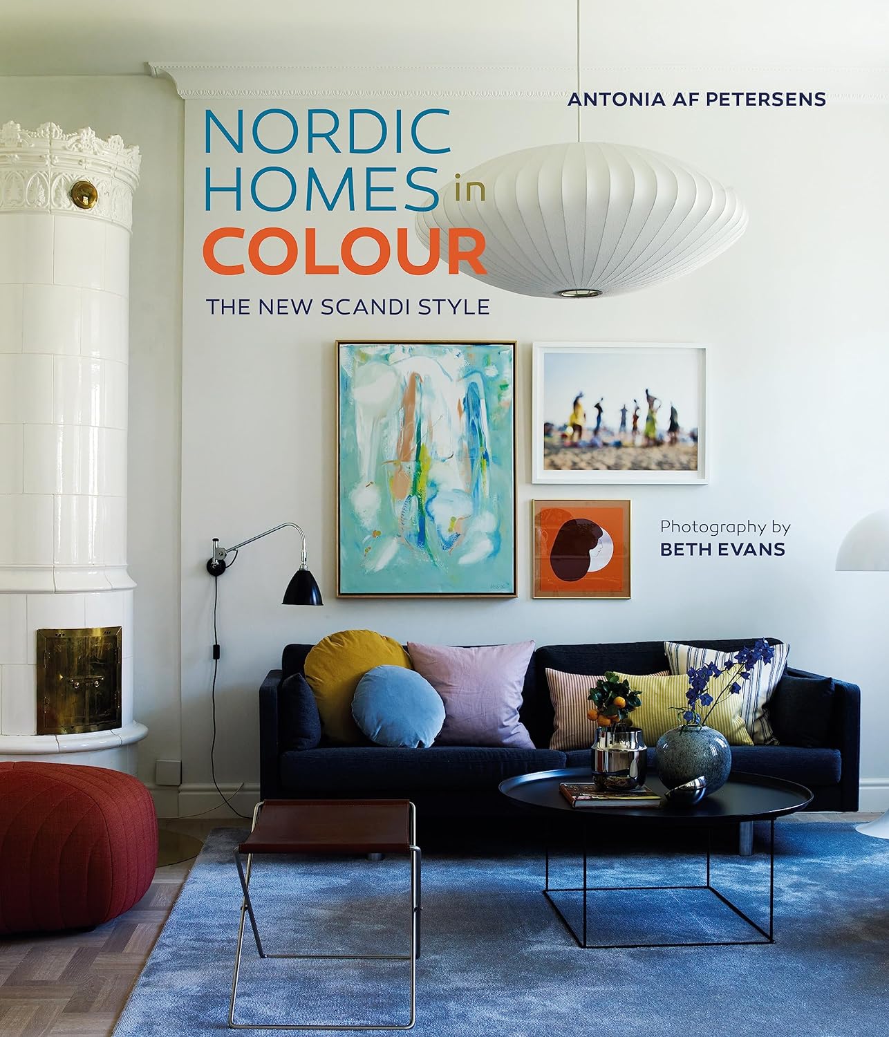 A modern living room with a dark sofa, vibrant cushions, abstract wall art, and round coffee table glows under a white pendant lamp on the cover of Nordic Homes in Colour - The New Scandi Style by Antonia af Petersen.
