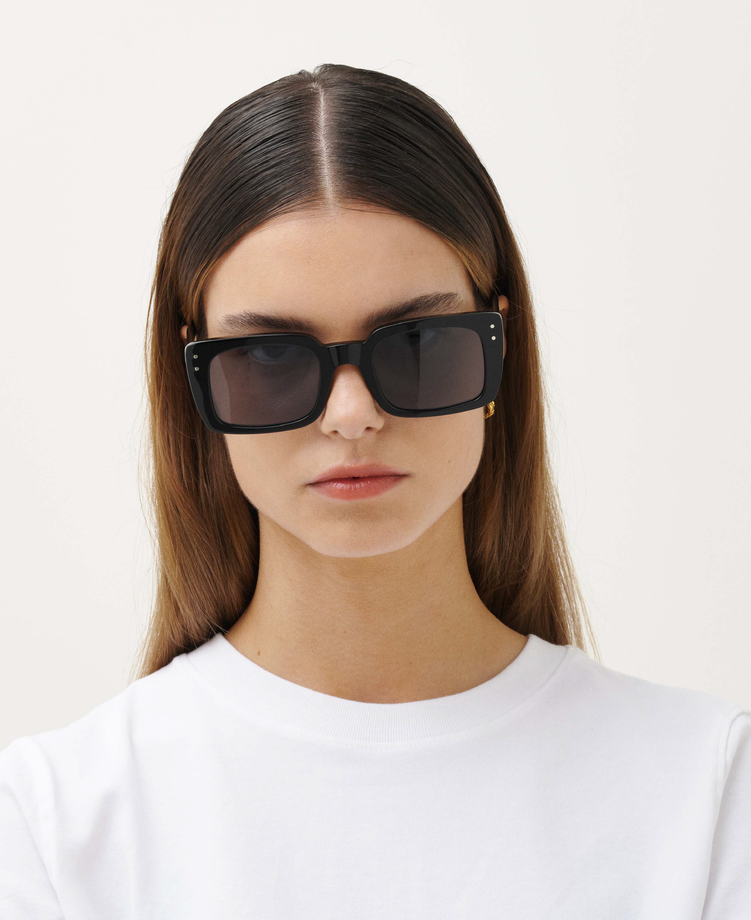 A woman with straight brown hair wears oversized MESSYWEEKEND Sunglasses Anna in Black with grey lenses and an acetate frame, paired with a plain white crew neck shirt, facing forward against a light background.
