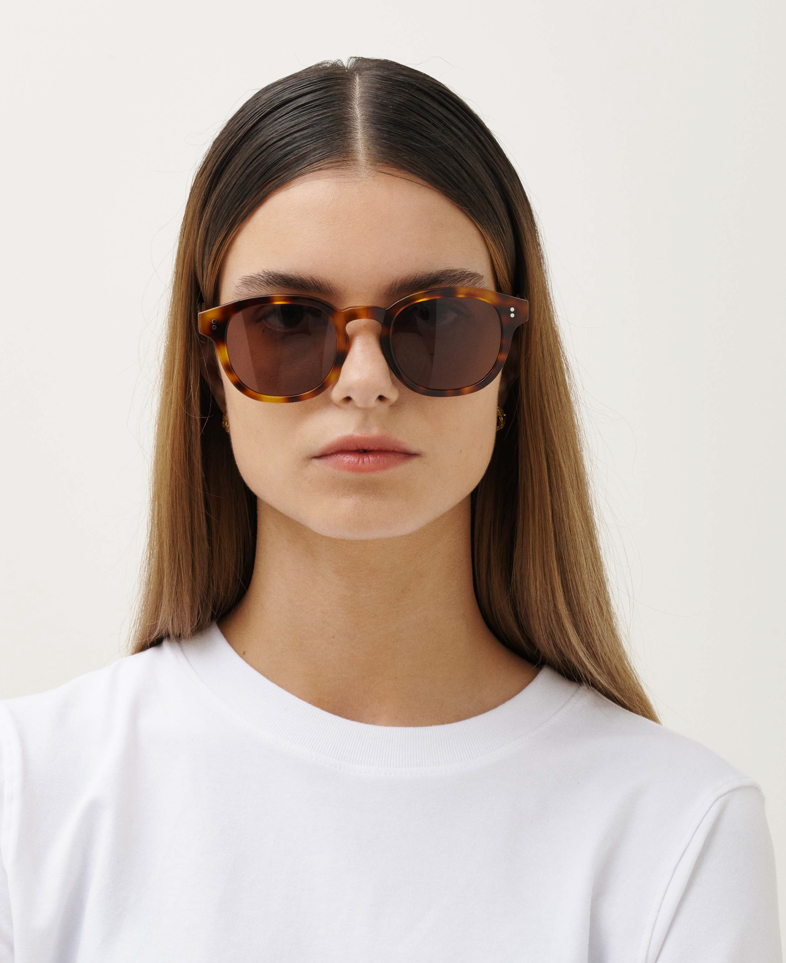 A woman with straight brown hair wears MESSYWEEKEND’s Sunglasses Billie in Tortoise with brown lenses and a plain white t-shirt, set against a light background.