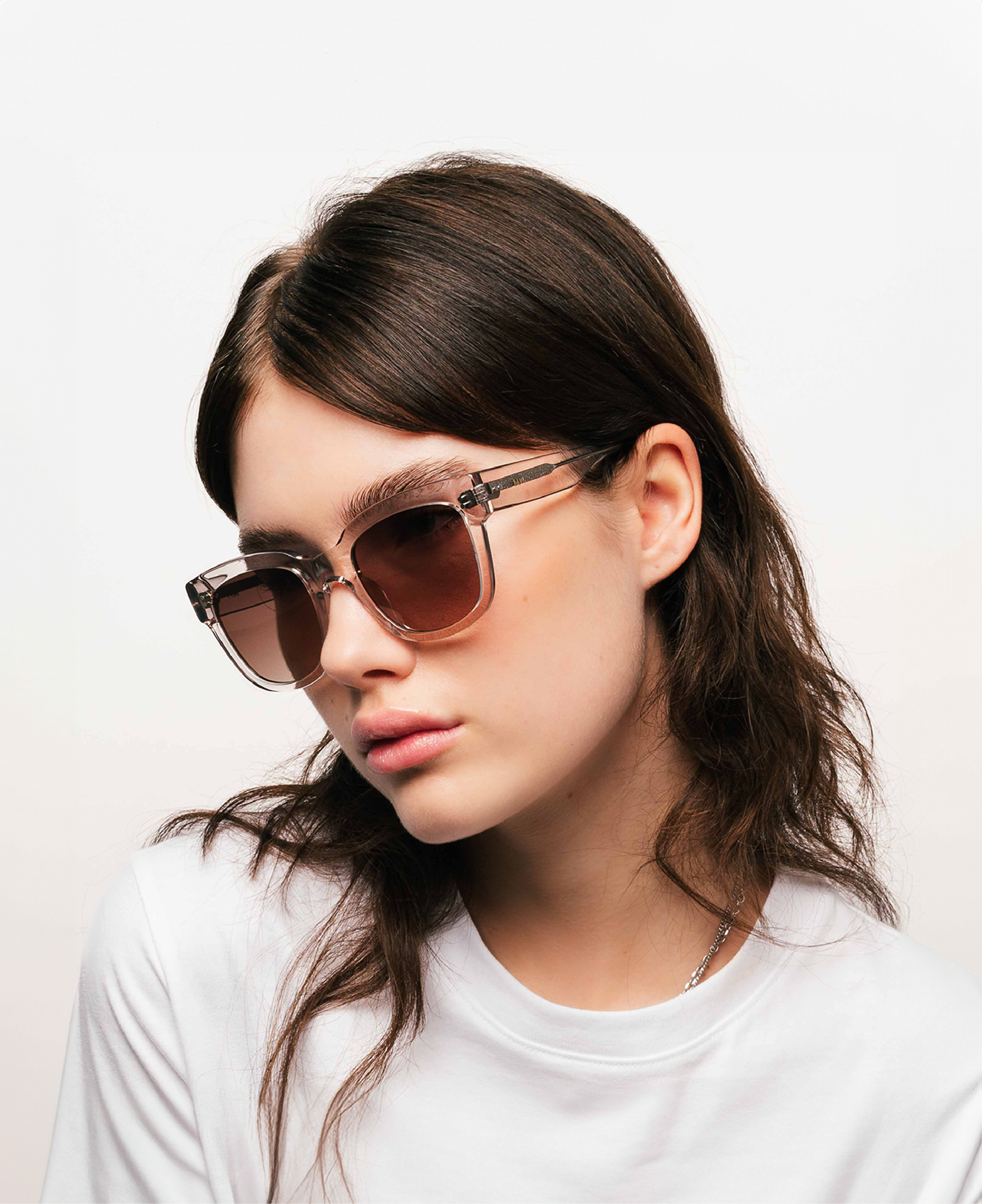 A woman with long brown hair wears MESSYWEEKEND Sunglasses Liv in Rose Clear with brown lenses—handcrafted acetate frames—paired with a white shirt, set against a plain light background.