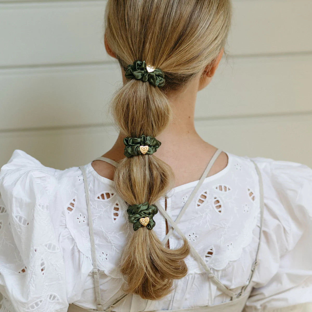 A woman with blonde hair in a bubble ponytail, styled with three ByEloise Silk Scrunchie Gold Heart in Olive Green. She wears a white embroidered blouse and is seen from behind against a light background.