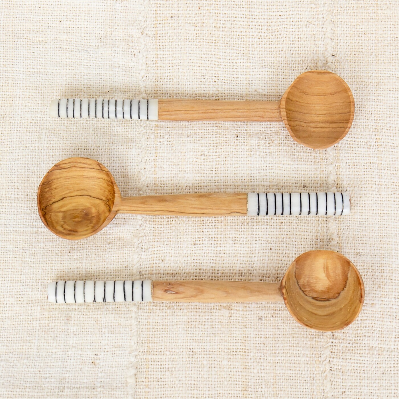 Three Striped Wooden Spoons by A World of Craft, each with a round bowl and white handle with black stripes, are arranged on woven beige fabric—showcasing the artistry of fair trade craftsmanship.