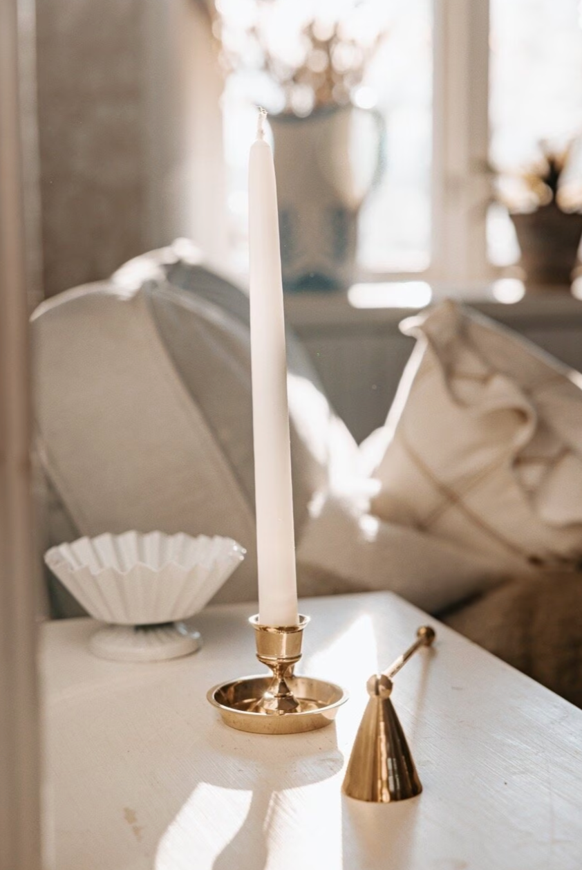 A white taper candle in a genuine brass holder sits on a light table beside the Strömshaga Candle Snuffer in Brass and a white ceramic bowl, with a sofa and vase visible in the background.