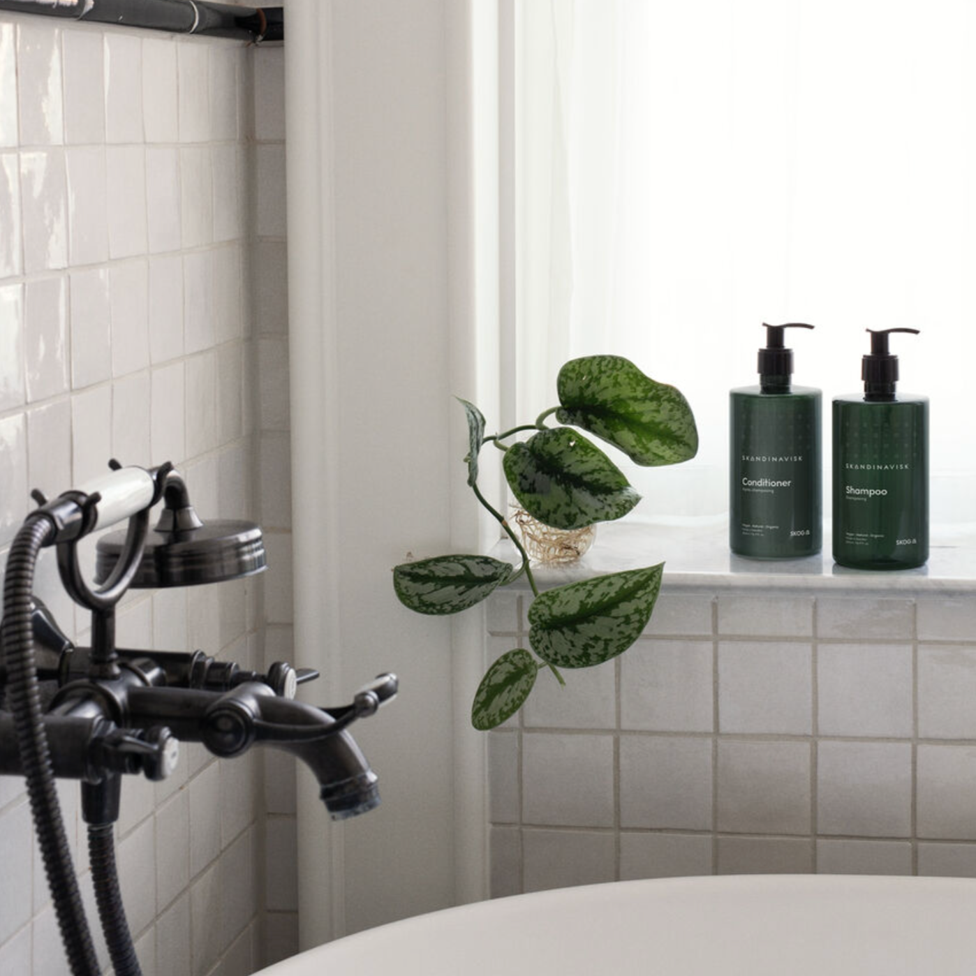 A bathtub with a black faucet, a leafy plant, and two green bottles of Skandinavisk Shampoo Skog 500ml with a vegan organic formula sit on a tiled ledge by a window.