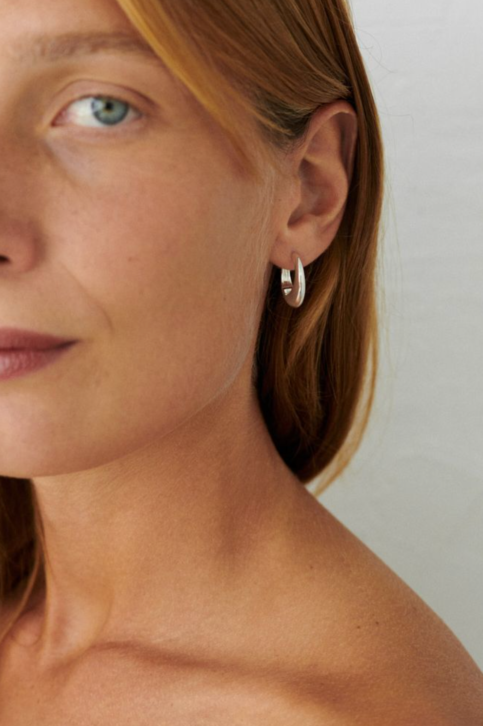 Close-up of a woman with fair skin and straight red hair wearing Pernille Corydon’s Globe Huggies in Gold, a small hoop earring by the Danish brand, with a neutral expression and bare shoulders for a timeless jewelry look.
