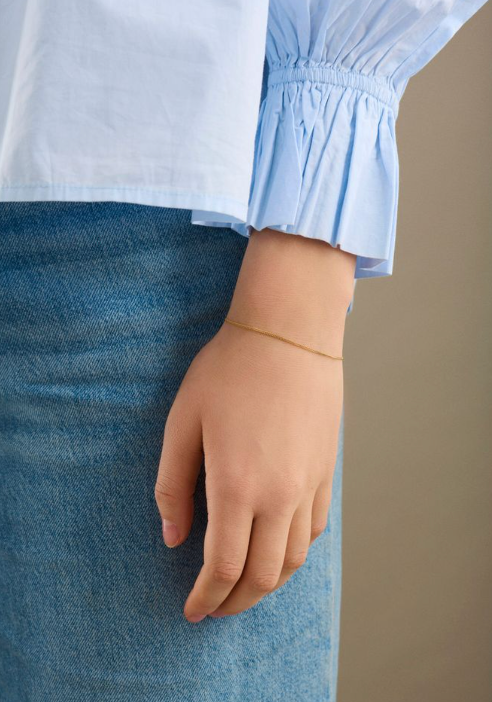 Wearing a light blue, long-sleeved blouse and blue denim skirt, they accessorize with the Pernille Corydon EA Bracelet in Gold—a subtle personal touch. Their hand is relaxed and resting by their side.