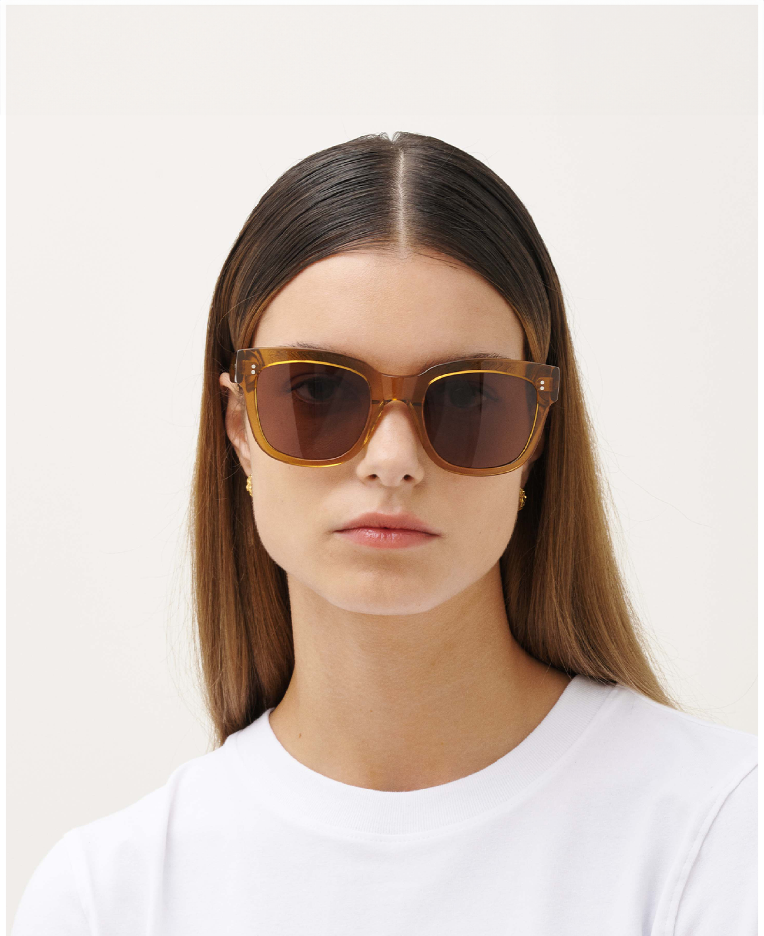 A woman with straight brown hair, wearing MESSYWEEKEND Sunglasses Liv in Coffee Brown with brown lenses and handcrafted acetate frames, faces forward in a white crew-neck shirt against a plain light background.