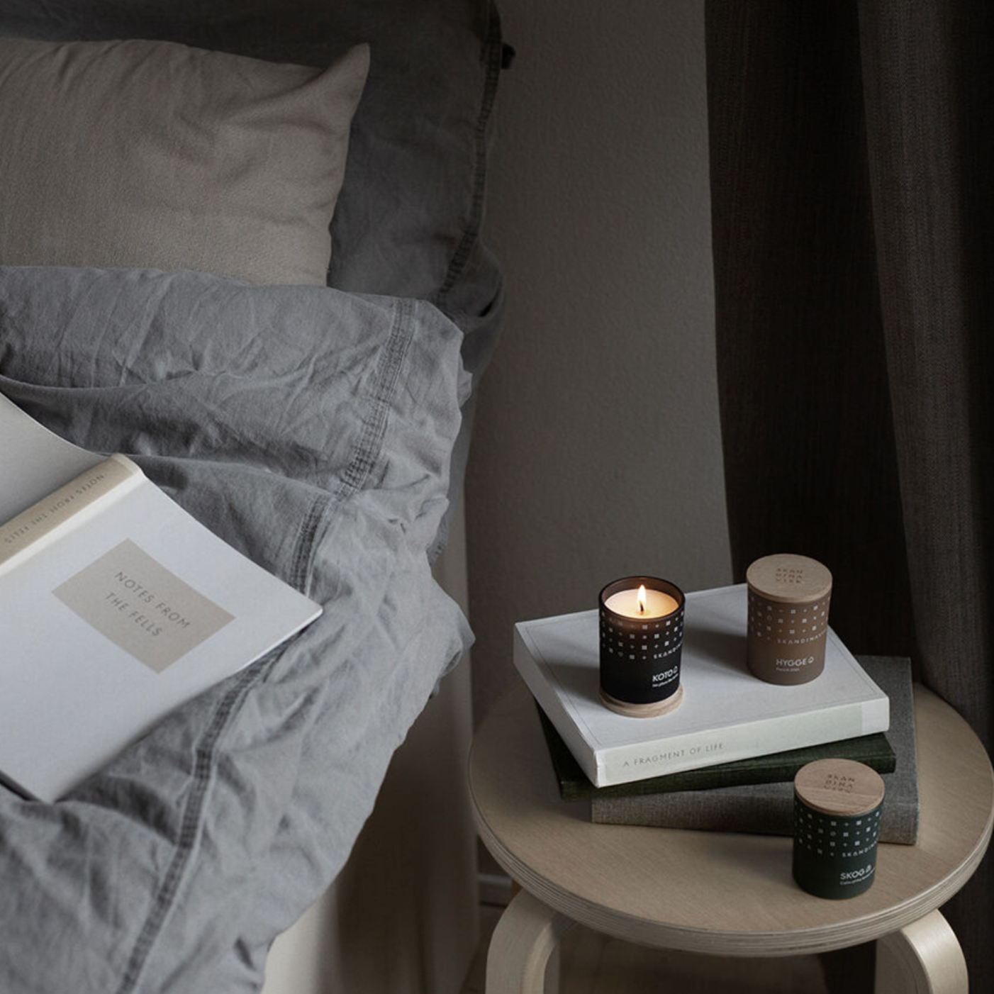 A neatly made bed with grey bedding stands next to a round table holding a lit mini candle from the Skandinavisk Scented Candle Hibernate Mini Giftset w. Skog, Koto & Hygge, along with two books, in a softly lit room.