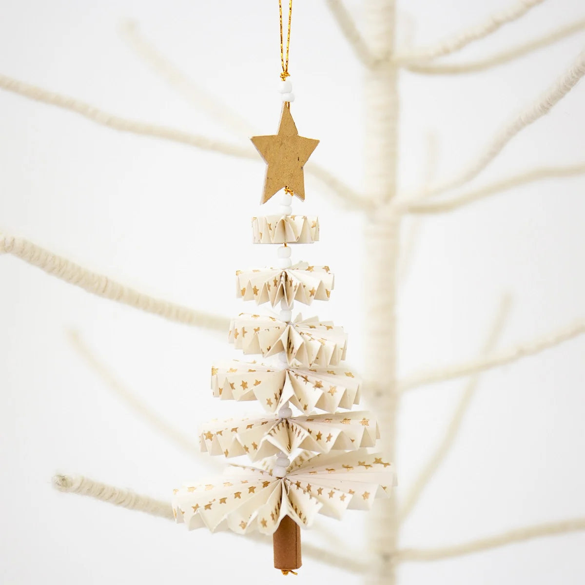 The Fold Fir Tree Christmas Ornament in white and gold by A World of Craft hangs from a bare white branch, bringing an eco-friendly, handmade holiday touch to your decor with its star-topped design.