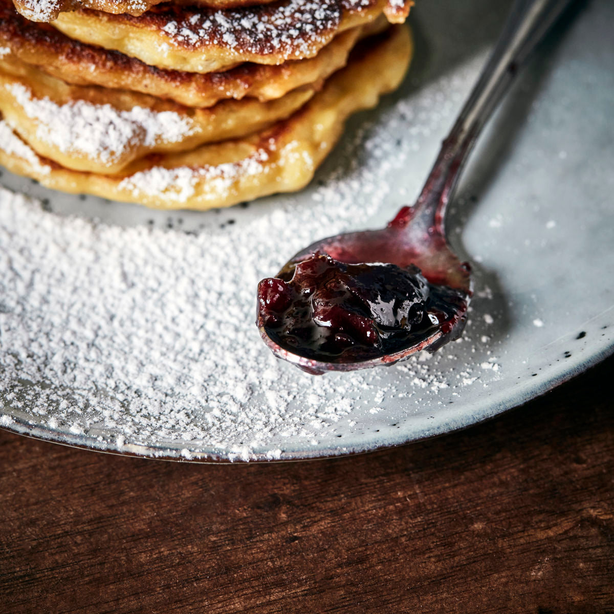 A stack of pancakes dusted with powdered sugar is served with a spoonful of Nicolas Vahé Jam Seasonal - Black Cherry & Mulled Wine.