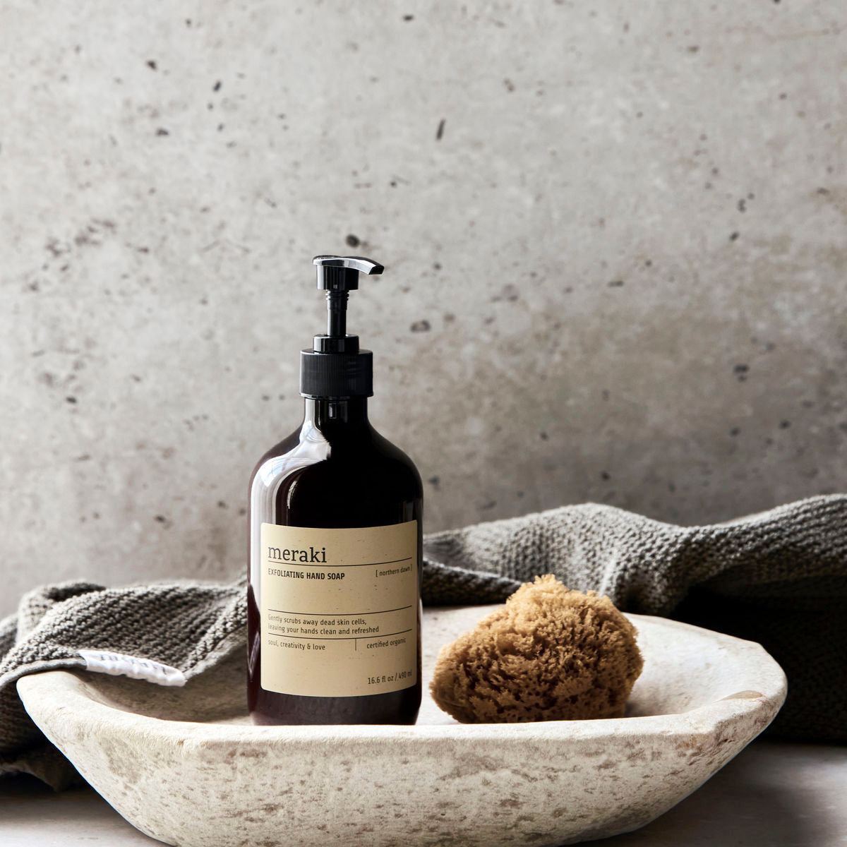 A brown Meraki “Hand Soap Exfoliating Organic, Northern Dawn” pump bottle rests in a stone dish with a sponge and towel on a neutral surface, set against a speckled stone background—perfect for displaying natural elegance.