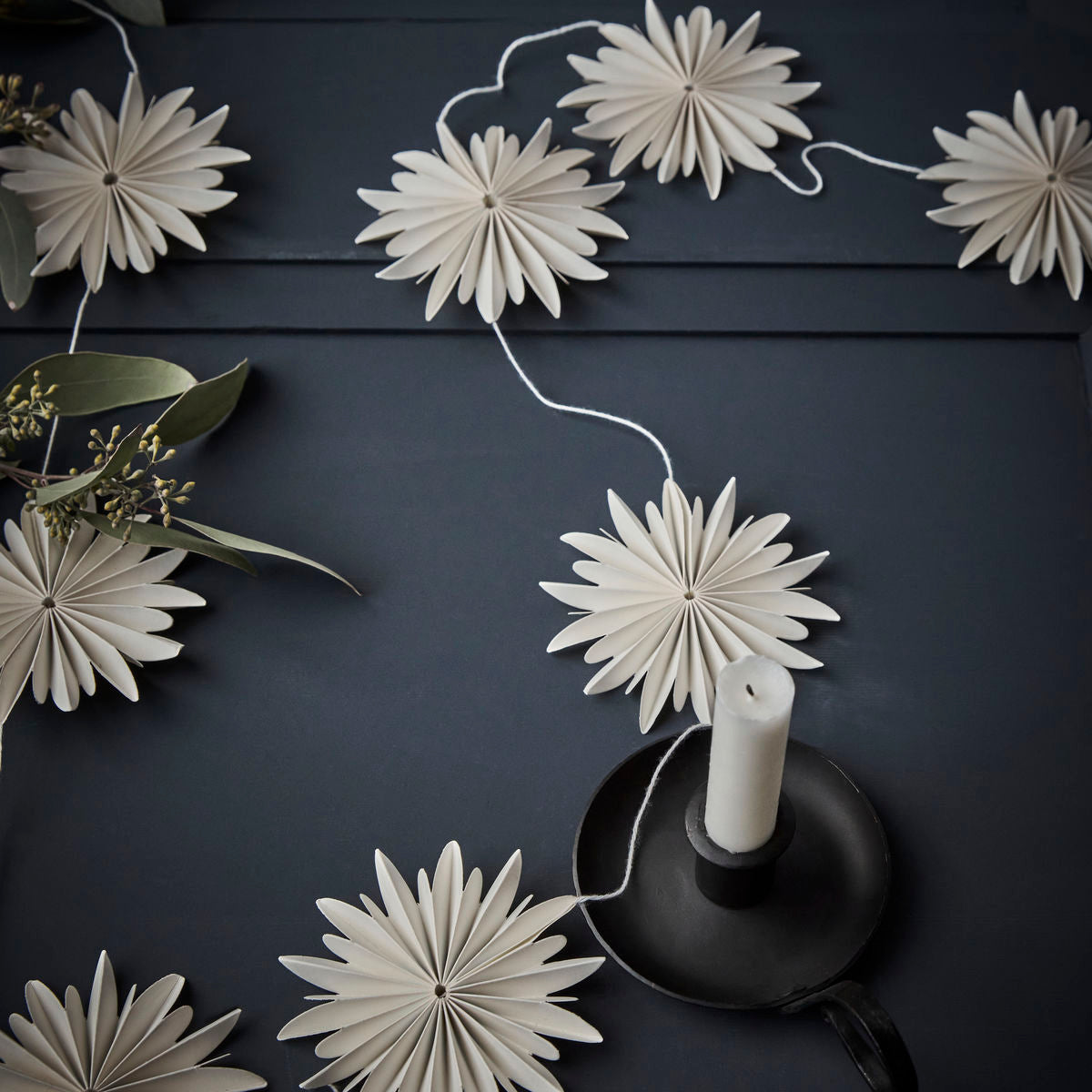 The Garland Girla Stars in Paper, White by House Doctor is arranged on a dark surface with a black candle holder and green foliage for a handmade decorative touch.