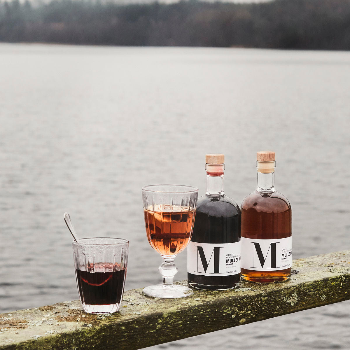 Two bottles and two glasses of Nicolas Vahé Mulled Wine Extract - Apple, Elderflower & Spices, a festive Christmas drink, sit on a weathered wooden railing beside a calm lake with trees in the background.