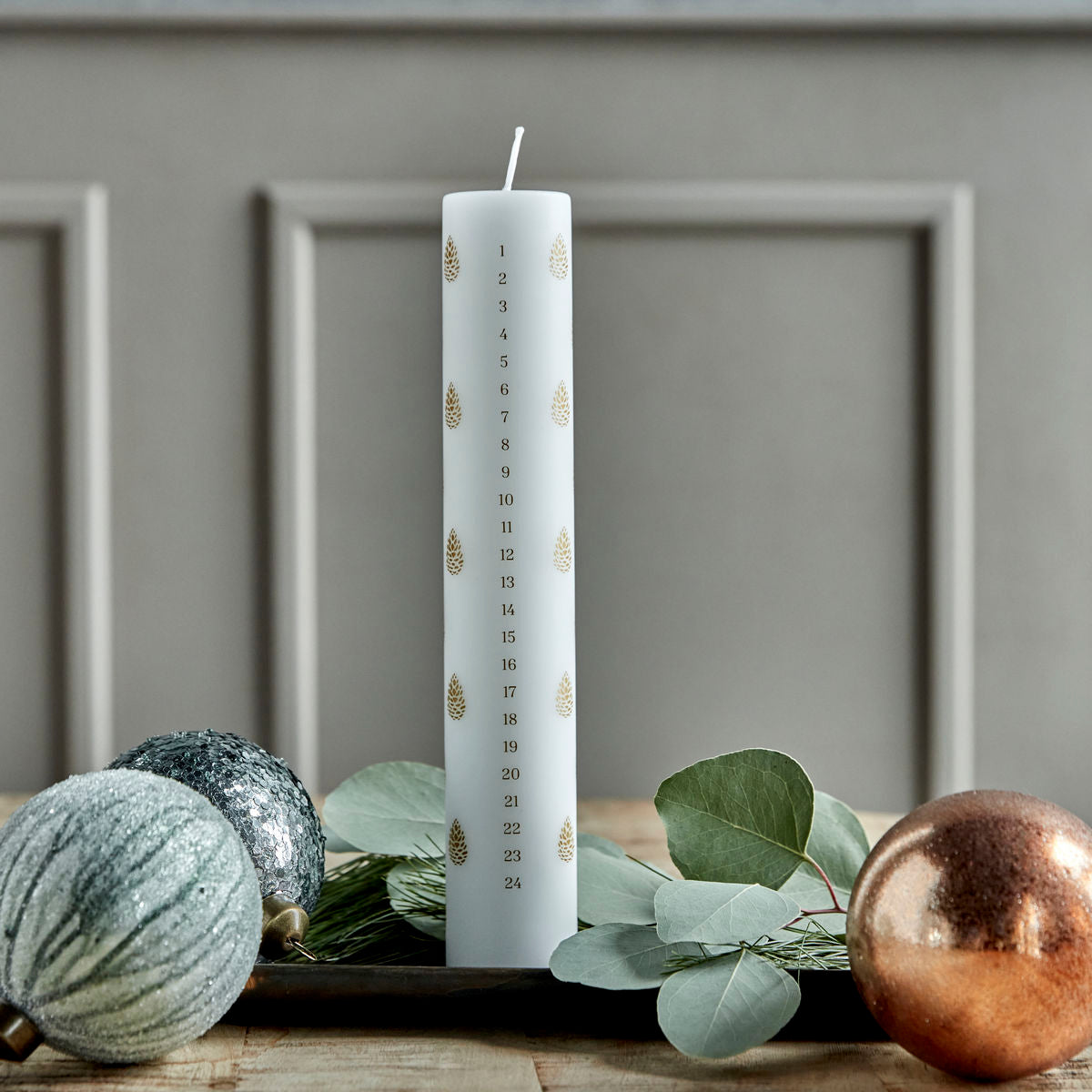 The House Doctor Calendar Candle 24 days of Christmas, Pinecones in White (4 cm), with gold pine cone designs and numbered days, sits on a wooden surface among ornaments and eucalyptus leaves for a festive countdown.