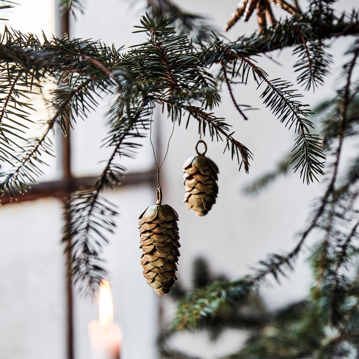 The Ornaments Pinecones in Iron, Set of 2 by House Doctor hang from a fir branch indoors, with a lit candle and blurred window behind them, adding an elegant touch to your Christmas décor.