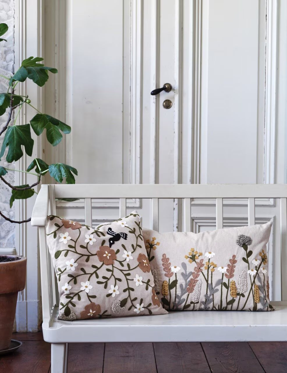 A white wooden bench with two Strömshaga Elodie Embroidered Beige with Flowers Cushion Covers (40x60 cm) is placed before a white paneled door, while a potted plant rests beside it on the wooden floor.