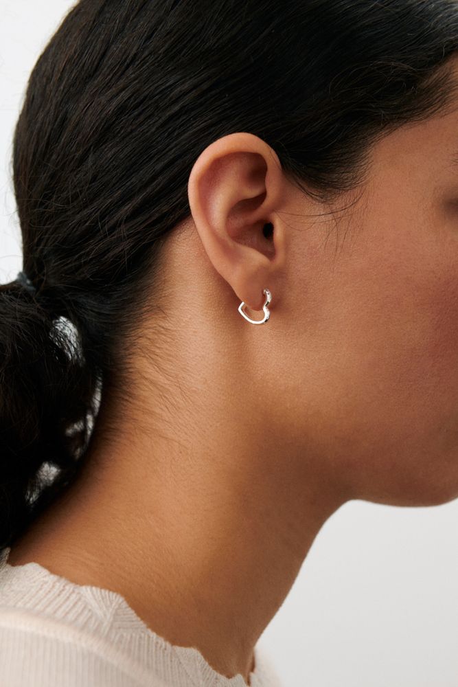 Close-up of a person with dark hair tied back, wearing Pernille Corydon’s Small Affection Hoops in Silver in their left ear. They are shown in profile and dressed in a light, textured top.