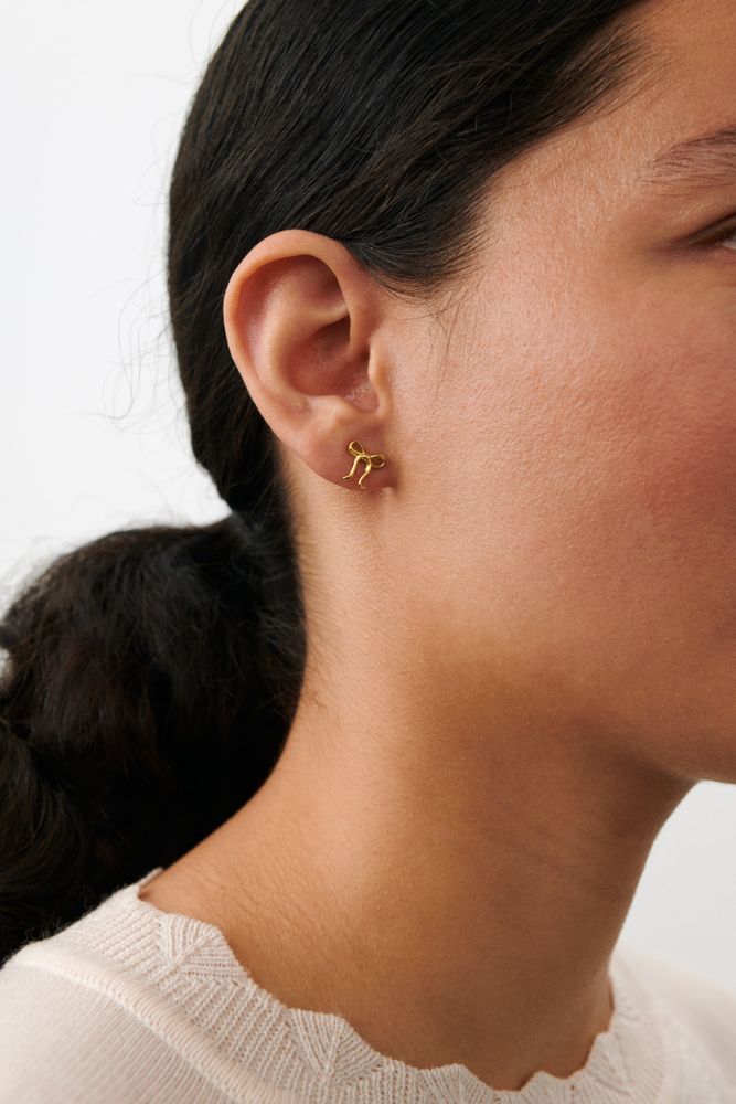 Close-up of a woman wearing Pernille Corydon’s Bow Stud Earrings in Gold, her dark hair pulled back and a light top against a white background—an elegant, feminine accessory.