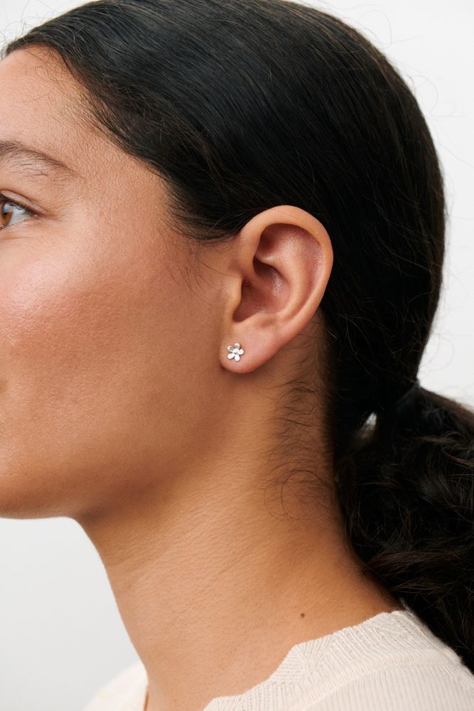 Close-up of a woman with dark hair in a low ponytail, wearing a cream-colored top and Pernille Corydon's Wild Poppy Stud Earrings in Silver in her left ear, against a plain light background.