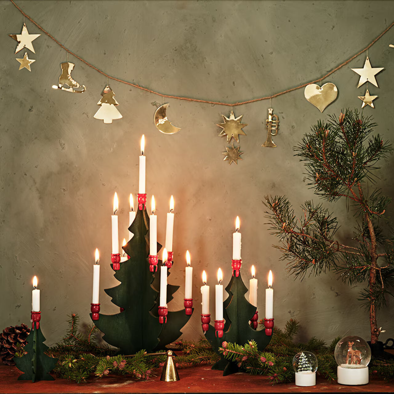 A festive scene with Strömshaga's 2.7m Garland with Ornaments in Brass, lit candles, pine branches, pinecones, classic Christmas motifs, gold accents, and a snow globe arranged on a wooden surface.