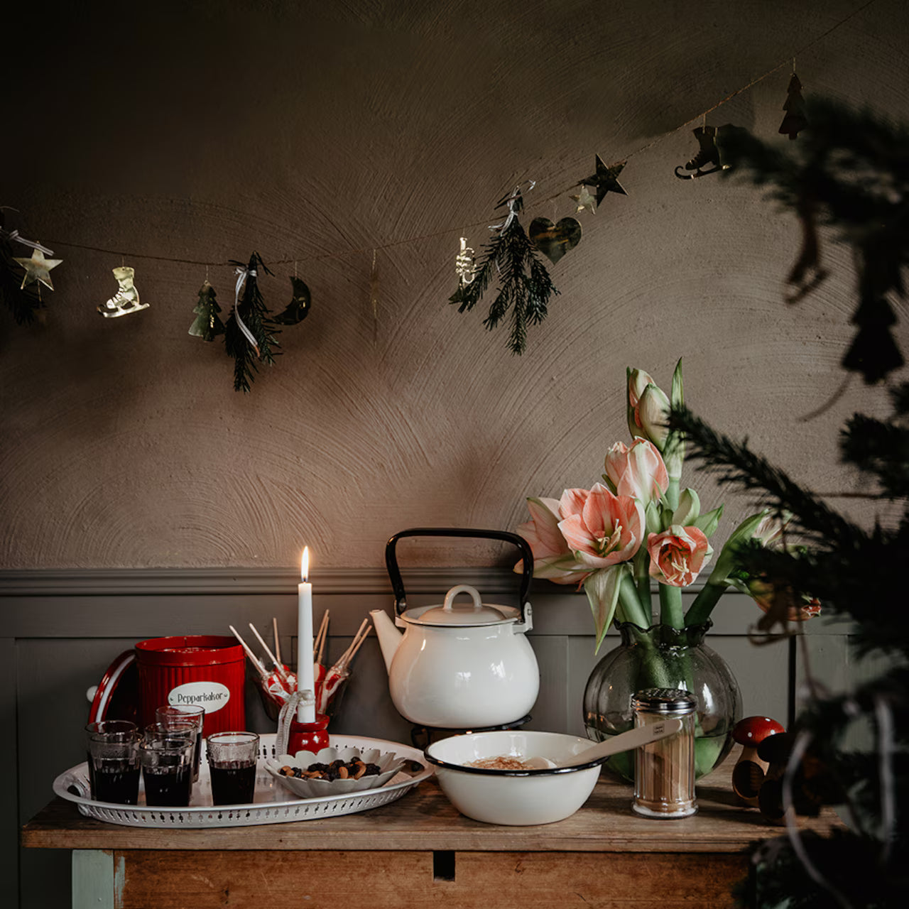 A festive table displays a white teapot, bowls, drinks on a tray, a lit candle, and flowers in a vase beneath Strömshaga's 2.7m Garland with Ornaments in Brass hanging against a textured wall.