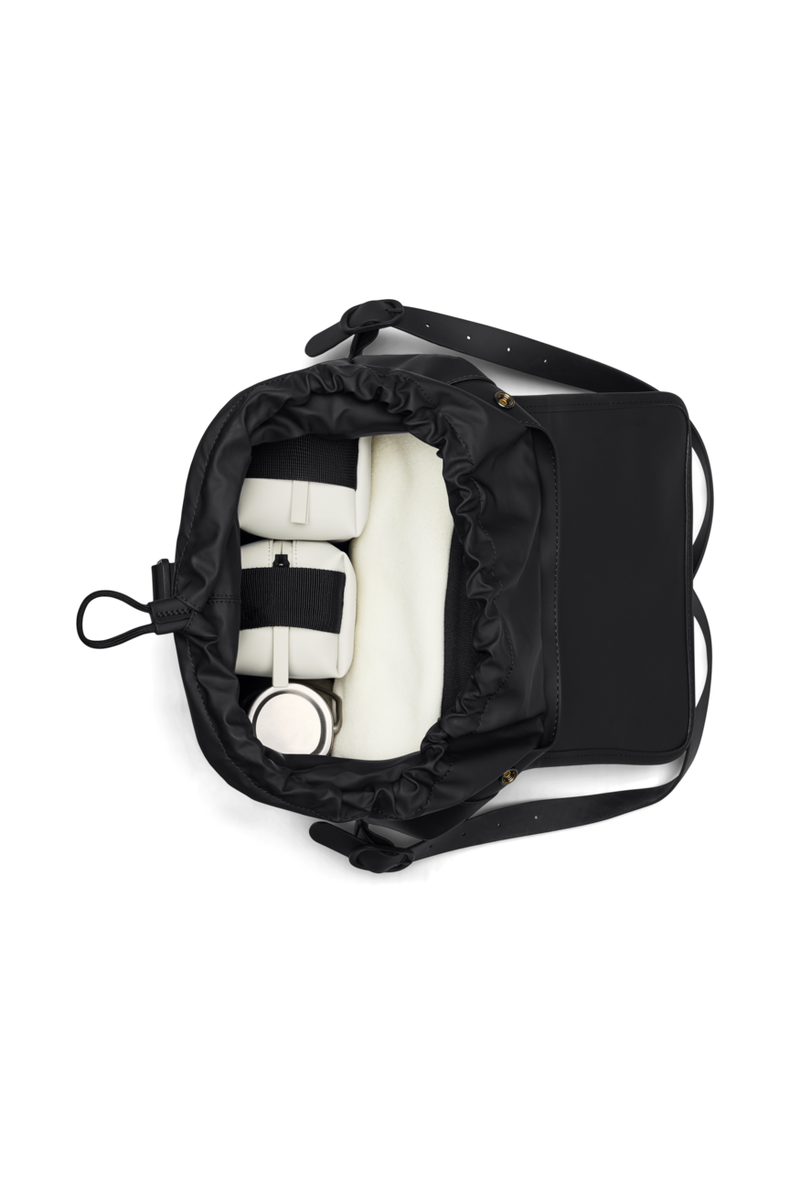 A top-down view shows the Rains Bucket Backpack in Black by Rains, crafted from PU fabric with a drawstring opening, holding two white camera lenses and a white camera.