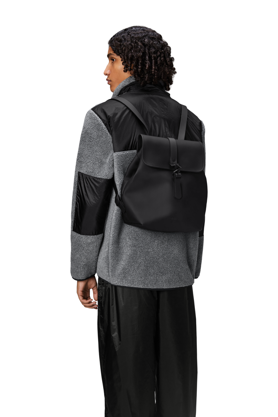 A person in a two-tone black and gray jacket and black pants, seen from behind, carries the Rains Bucket Backpack in Black by Rains—a weatherproof bag featuring a flap closure.