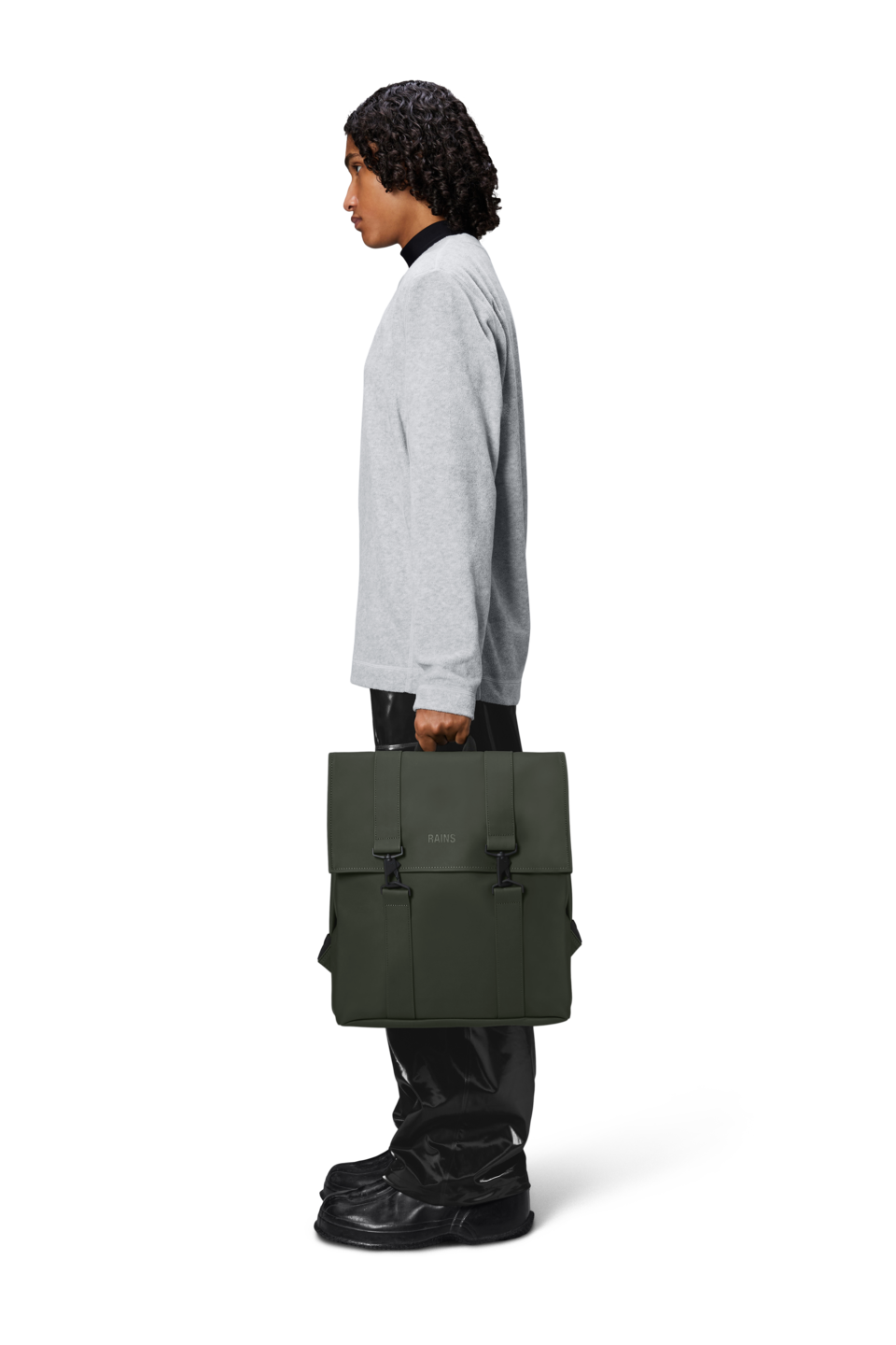 Person in profile wearing a gray sweatshirt and black pants, holding the Rains MSN Bag in Green—an urban essential from Rains, featuring dual buckles and a practical laptop pocket—against a plain white background.