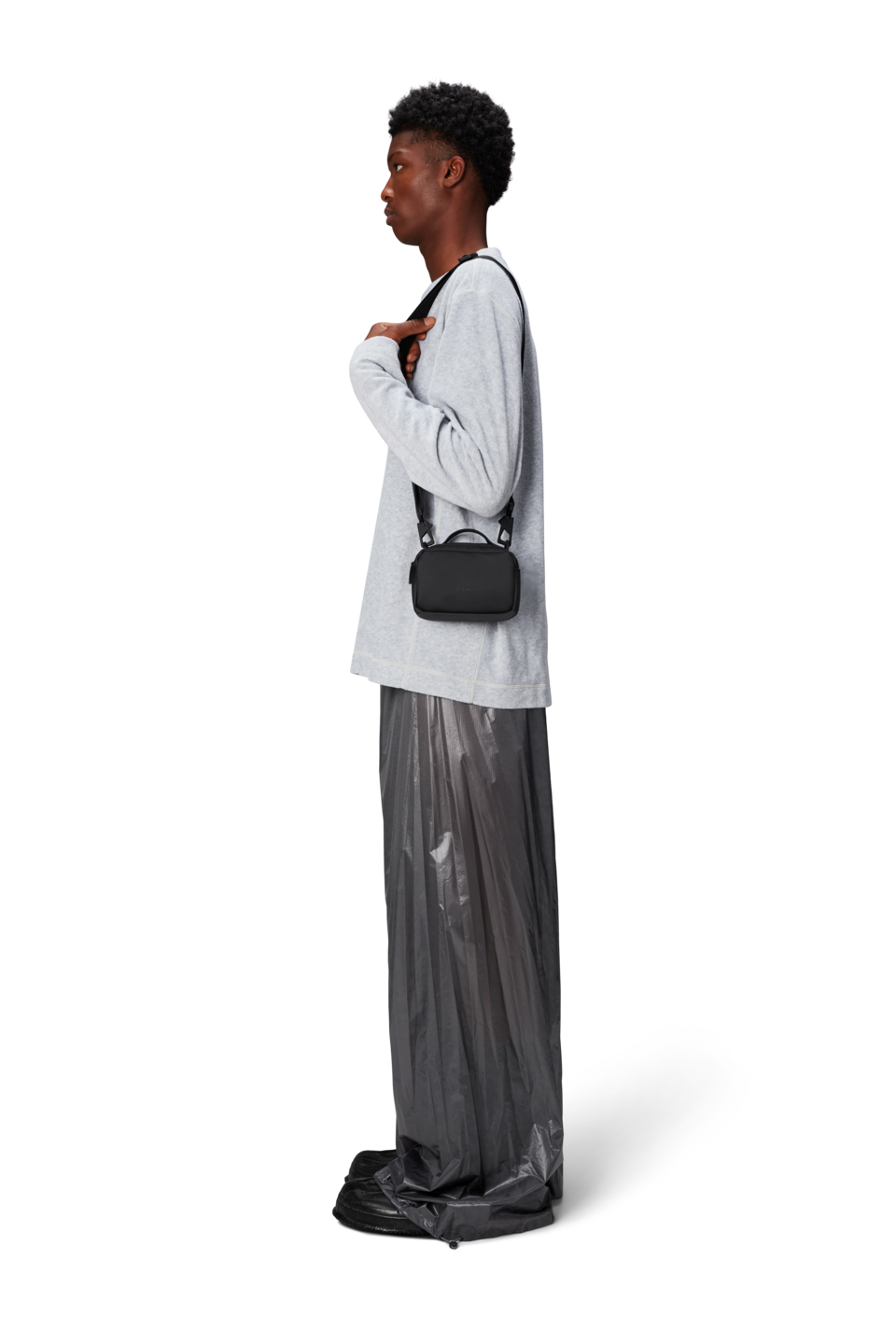 A person in profile wears a light gray long-sleeve shirt, shiny loose gray pants, black shoes, and carries the Rains Box Bag Micro in Black by Rains—a small waterproof bag ideal for daily essentials.