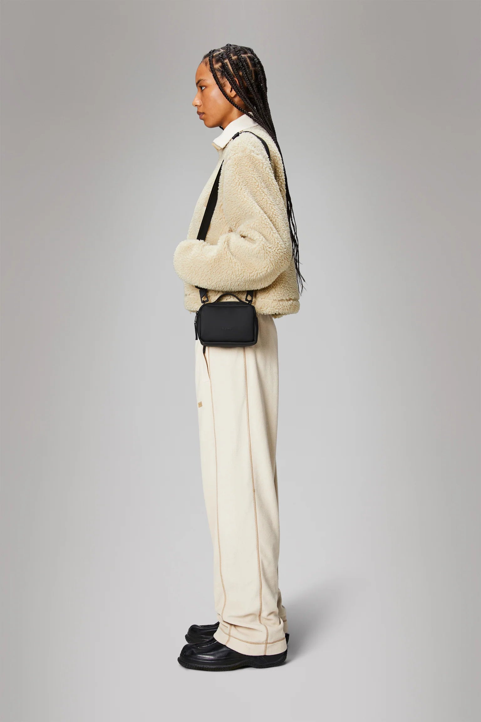 A person with long braided hair stands in profile against a plain gray background, wearing a beige sherpa jacket, cream pants, black shoes, and carrying the Rains Box Bag Micro in Black by Rains for daily essentials.
