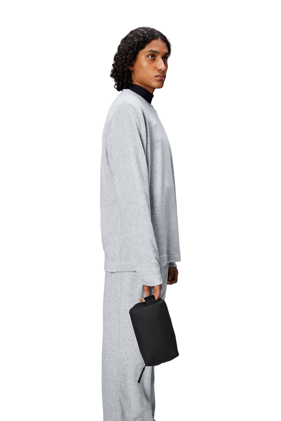 A person with curly hair wears a loose grey sweatshirt and matching pants, holding the Rains Wash Bag Small in Black by their side. The background is plain white.