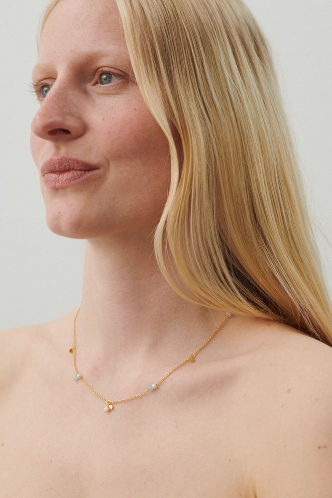 A woman with long blonde hair wears the Pernille Corydon Afterglow Sea Necklace in silver, adorned with freshwater pearls and blue agate, evoking natural beauty against a plain light background.