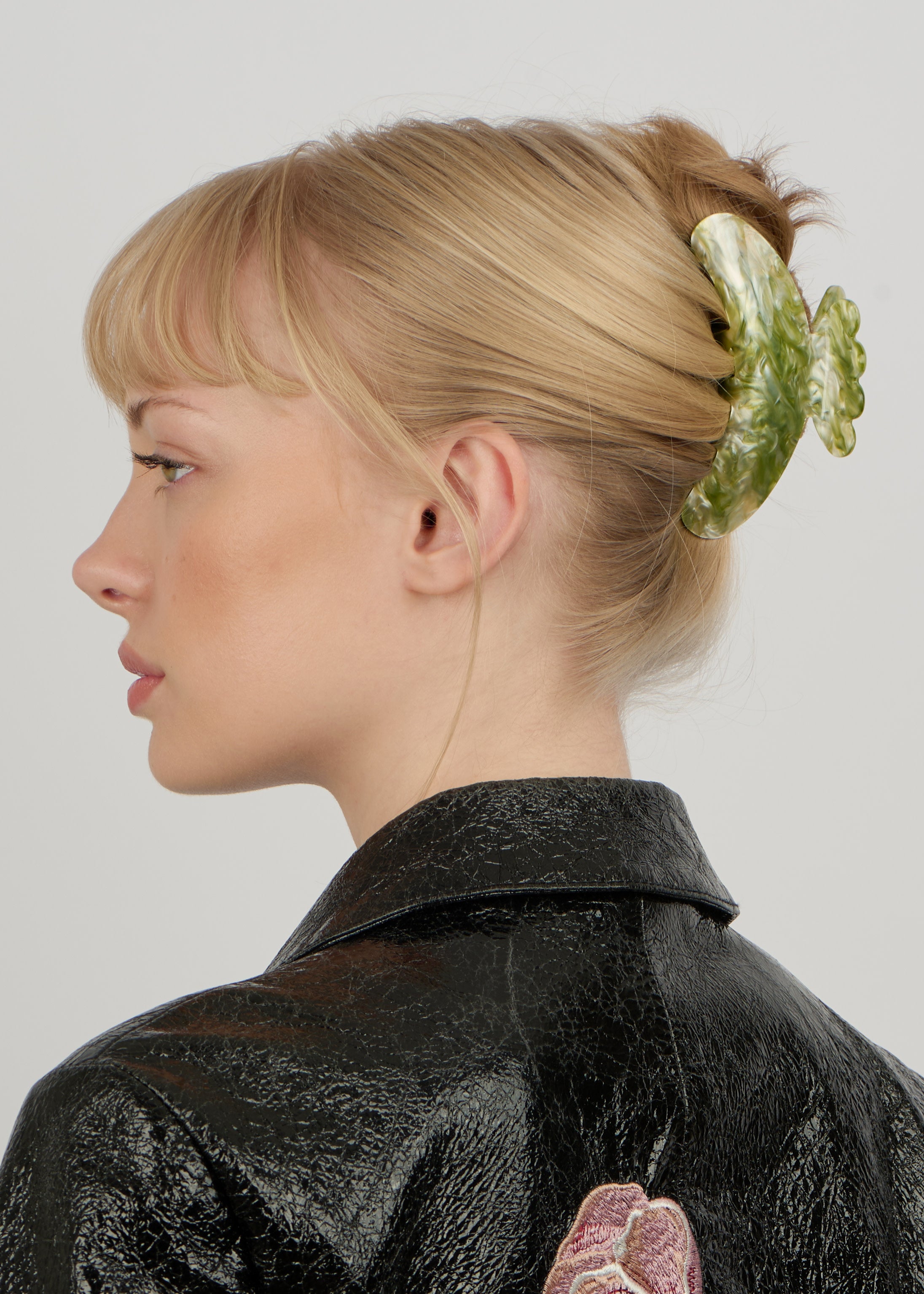A person with blonde hair styled in a twist, secured with the Sui Ava Helle Matcha Big Hair Claw Clip in Cool Green, is shown in profile, wearing a textured black jacket featuring embroidered detailing on the back.