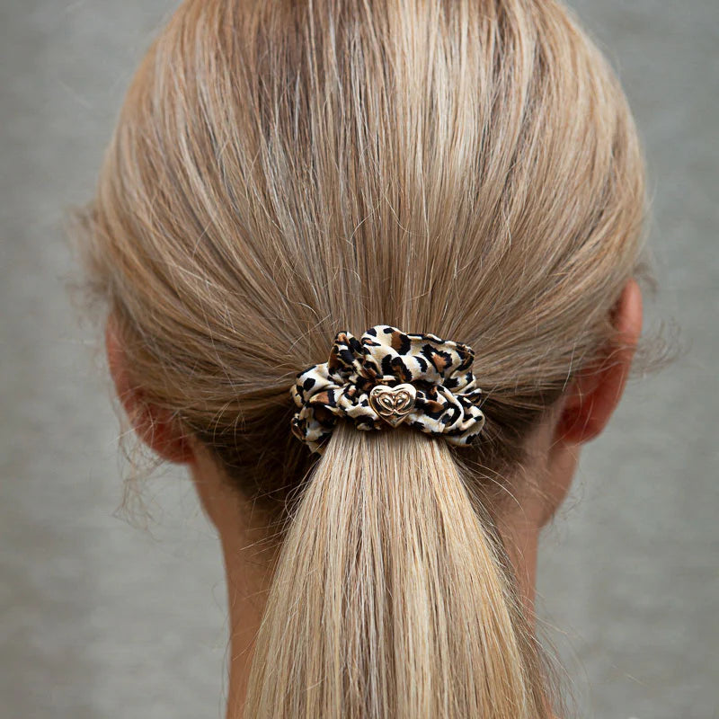 A person with blonde hair in a low ponytail, secured with a ByEloise Silk Scrunchie Gold Heart · Leopard, adorned with a small metallic gold heart charm.