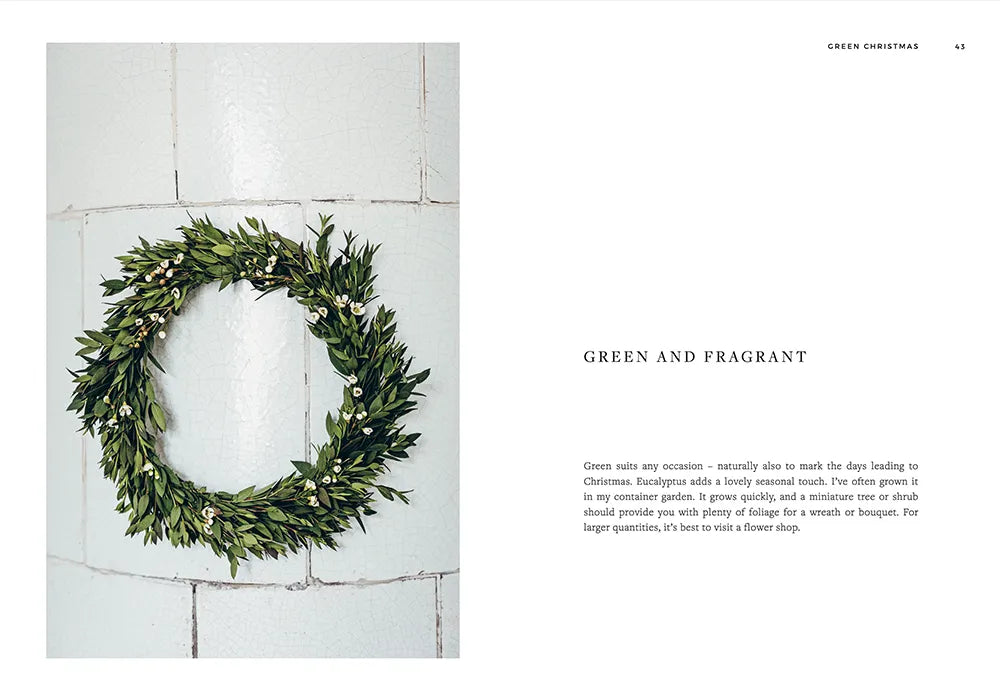 A Green Christmas - Wreaths & Floral Arrangements by Cozy Publishing is displayed on a white tiled wall, featuring a festive eucalyptus wreath next to a page highlighting greenery and Christmas floral decor ideas.