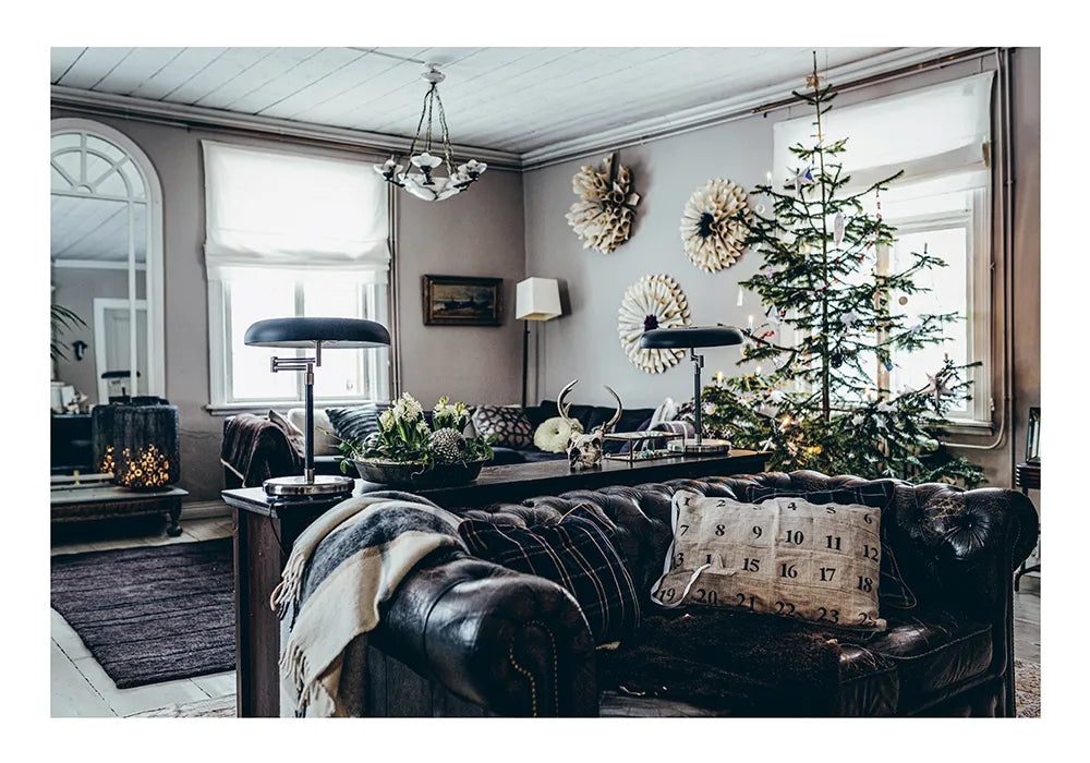A cozy living room with a leather sofa, festive pillows, throw blanket, and desk lamps features large windows and Green Christmas - Wreaths & Floral Arrangements by Cozy Publishing for a charming holiday touch.