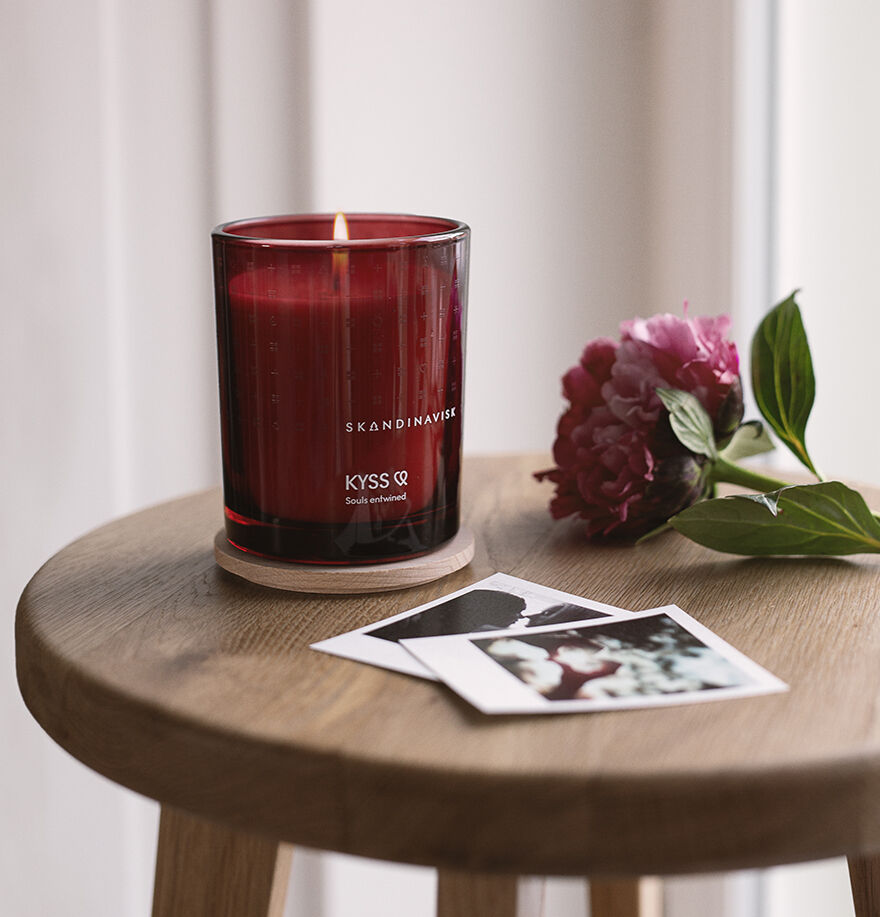 A Skandinavisk Scented Candle Kyss, 200 g, in a glass holder rests on a round wooden table beside a pink peony and two instant photos, illuminated by soft natural light from the window.