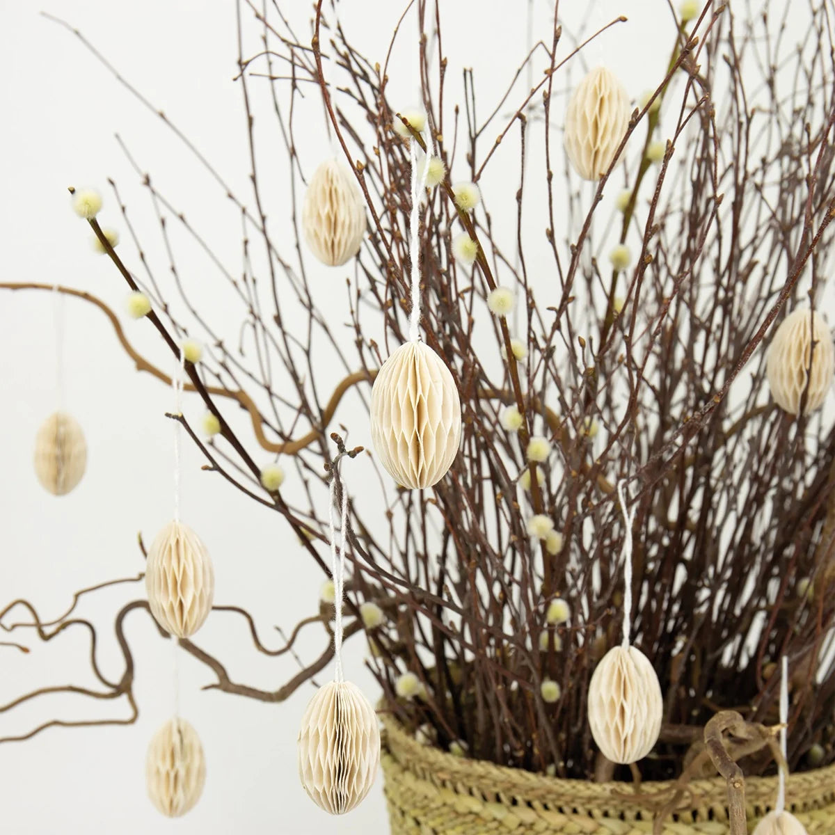 A woven basket with bare branches and pussy willow stems forms a charming Swedish Easter tree, adorned with beige Honeycomb Easter Egg Ornaments by A World of Craft. The scene is set against a plain white wall.