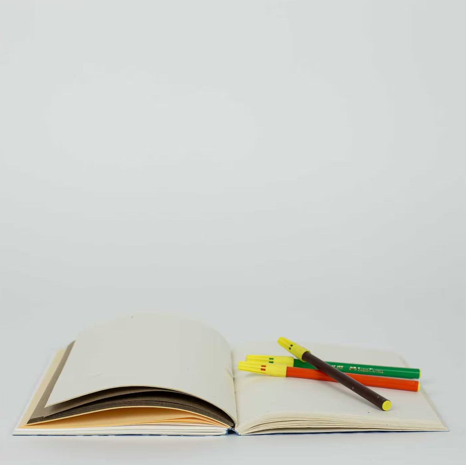 A World of Crafts blue A5 Dotted Notebook rests open on a white surface, topped with three colored pens in yellow, green, and brown.