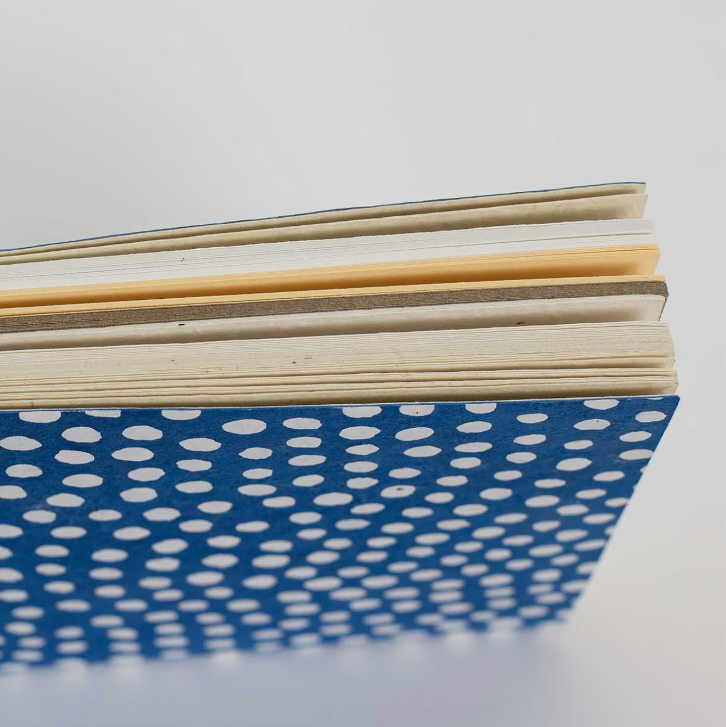 A close-up of the top edge of A World of Crafts Dotted Notebook, Blue, A5 highlights its handmade lokta paper, thick uneven pages, and blue cover with white polka dots.
